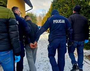 Na zdjęciu umundurowani policjanci prowadzą mężczyznę, który na ręce trzymane z tyłu ma założone kajdanki. Obok policjanci w ubraniach cywilnych.