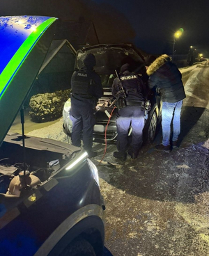 Na zdjęciu dwaj umundurowani policjanci i mężczyzna w ciemnej kurtce pochyleni nad pojazdem, jego maska jest otwarta.