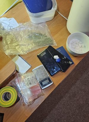 Na stole leżą przedmioty, plastikowe pudełko z niebieskim wieczkiem, worek z zielonym suszem, w białej miseczce biały proszek, dwa telefony komórkowe, waga, worek z małymi woreczkami strunowymi i zielona taśma izolacyjna.