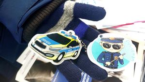 Ręka dziecka w wełnianej rękawiczce, na niej breloczek w kształcie policyjnego radiowozu i okrągła naklejka policjanta.
