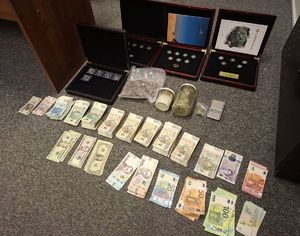 Na zdjęciu od góry klasery z okrągłymi przedmiotami przypominającymi monety, worek z suszem roślinnym, dwa słoiki z narkotykami, poniżej rozłożone pieniądze w polski walucie, niżej banknoty euro, funty i hrywny.