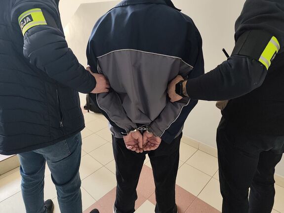 Na zdjęciu policjanci w ubiorach cywilnych, na przedramieniu mają odblaskowe opaski z napisem policja. Między nimi stoi mężczyzna, którzy ma założona na ręce trzymane z tyłu kajdanki.