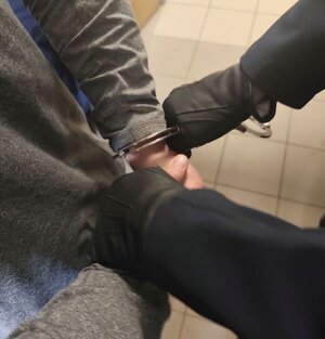 Na zdjęciu umundurowany policjant zakładający kajdanki na ręce z tyłu osobie zatrzymanej.